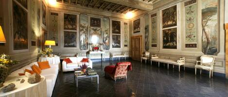 Una habitación amplia con techo alto, vigas ornamentales y suelo negro pulido. Hay varias zonas de estar con sofás y sillones tapizados en blanco y rojo. Las paredes están decoradas con cuadros enmarcados y paneles decorativos.