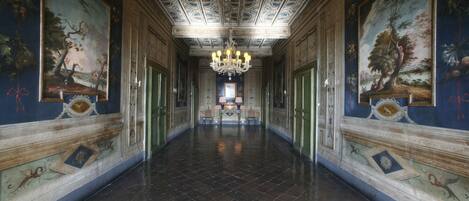 A grand hallway with a chandelier, ornate ceiling, and classical paintings.