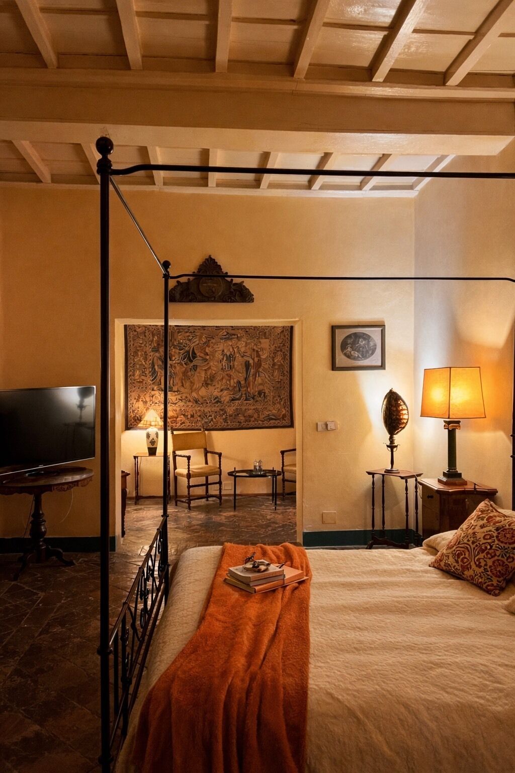 Ein Bett mit Baldachin, eine rote Tagesdecke, ein Flachbildfernseher, ein Gemälde und eine Lampe auf einem Nachttisch.