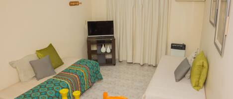 Una habitación pequeña y bien iluminada con dos camas individuales, un televisor, una mesa con saleros y pimenteros amarillos, y un equipo de aire acondicionado.