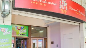 The exterior of the Patios del Mediterráneo apartment hotel with a storefront labeled 'Top Cel' for mobile services.