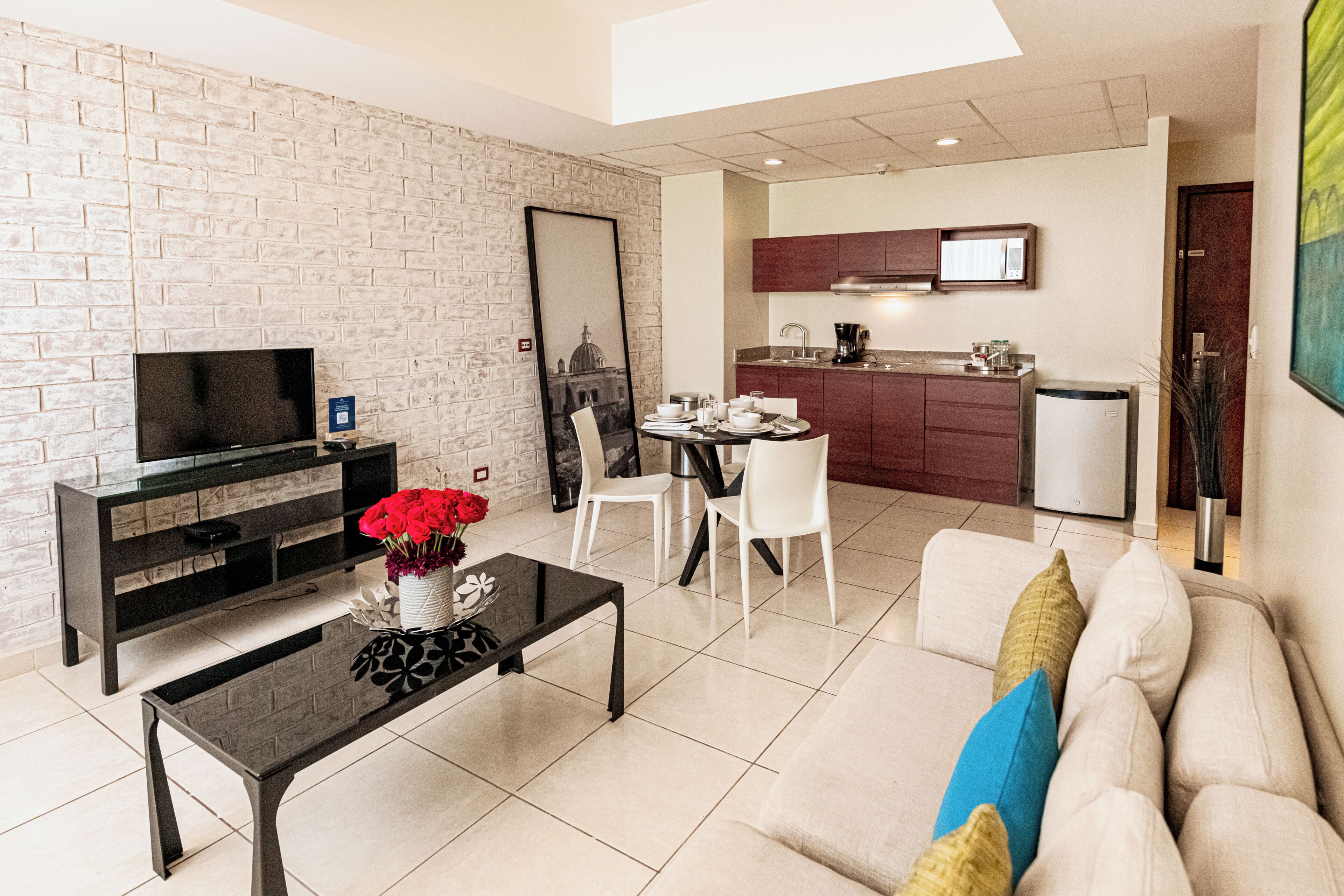 A modern living room with a TV, coffee table, and sofa, a dining area with chairs, and a kitchen with cabinets and a refrigerator.