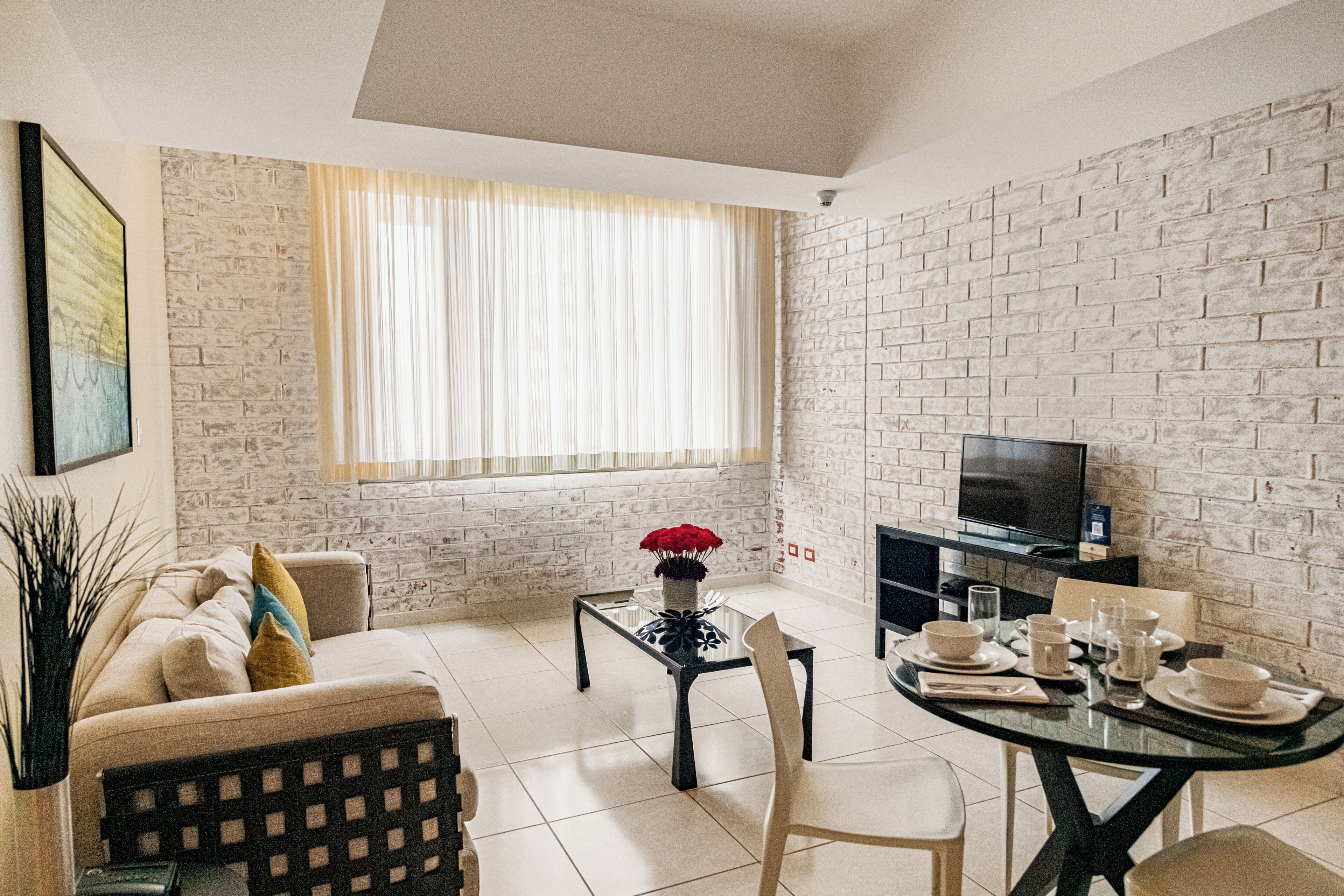 A living room with a sofa, a coffee table, a dining table set for four, a TV, and a brick wall.