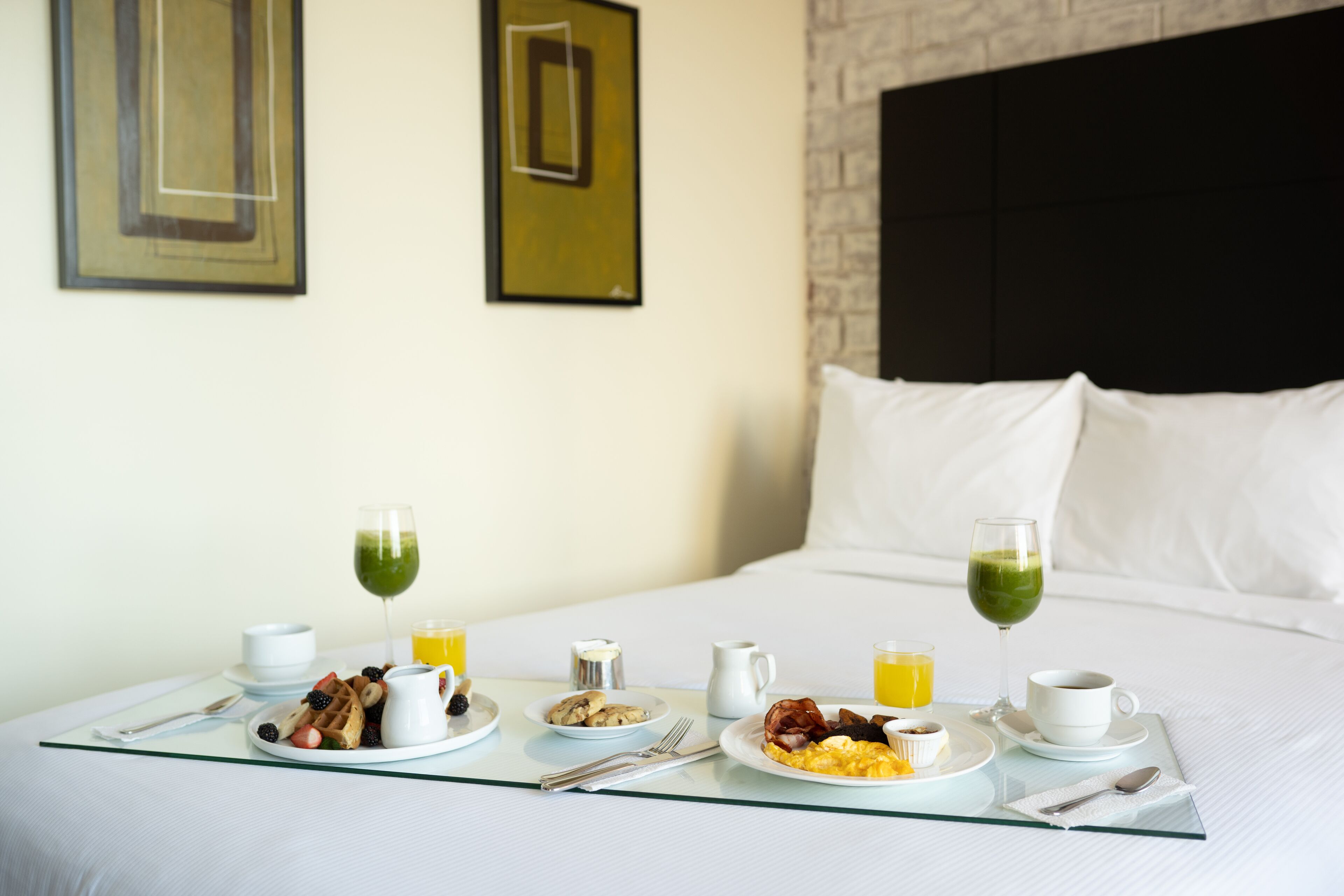 A neatly arranged breakfast table with plates of waffles, eggs, and fruit, accompanied by glasses of green juice and orange juice, set on a bed in a hotel room.