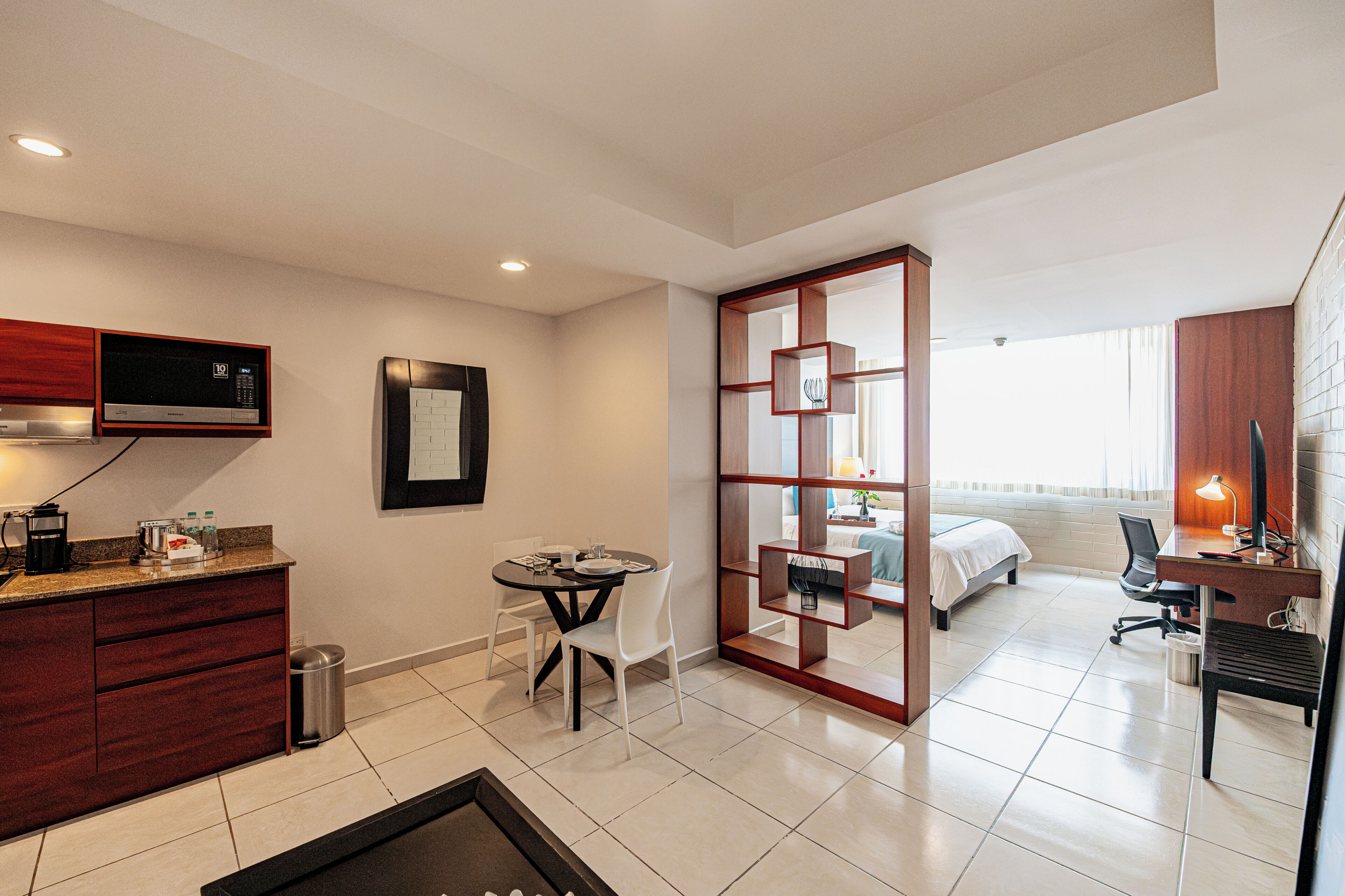A compact hotel room with a kitchenette, a dining area, a bed, a desk with a chair, and a large window.