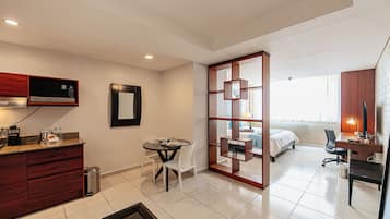 A compact hotel room with a kitchenette, a dining area, a bed, a desk with a chair, and a large window.