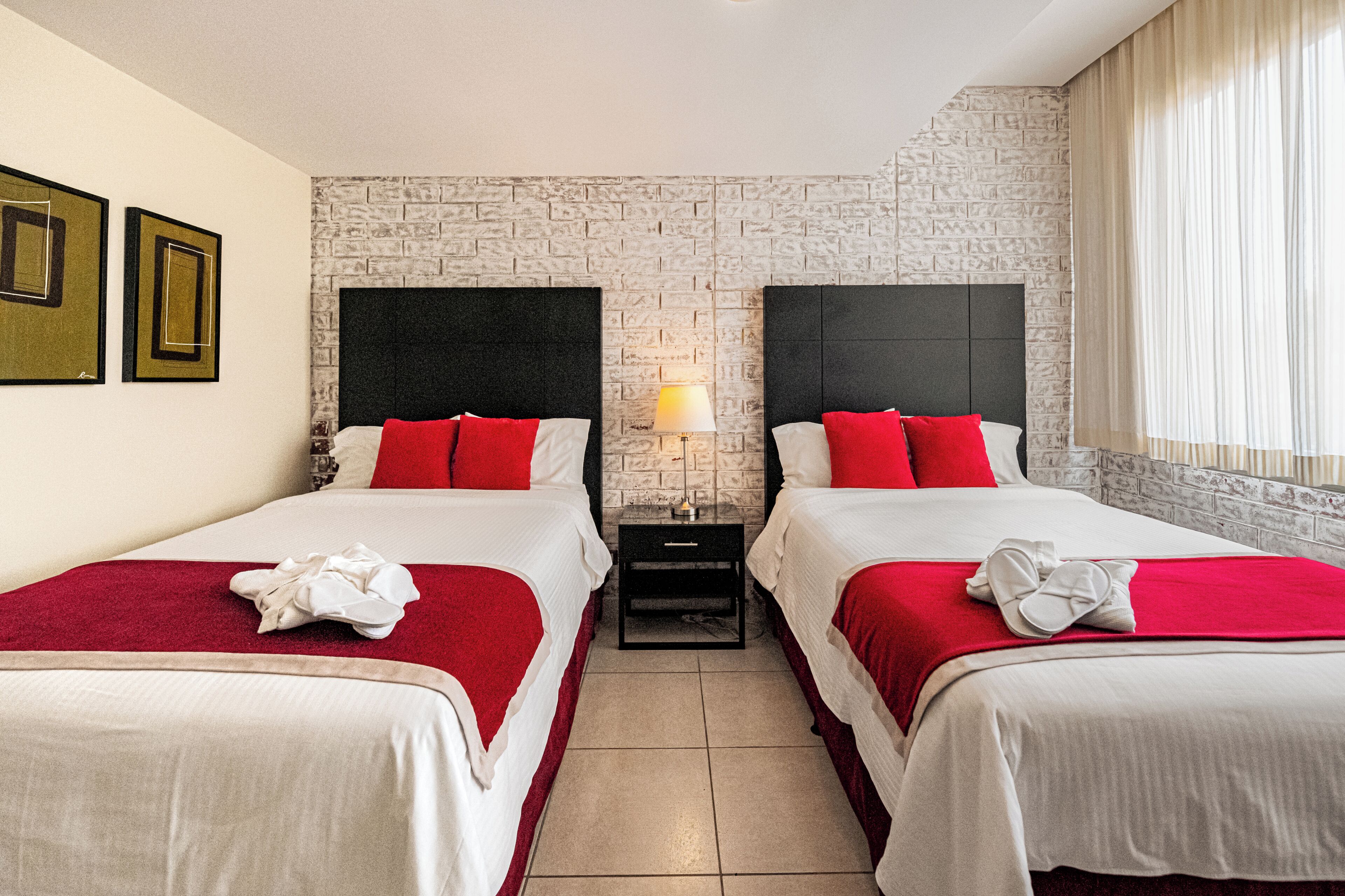 A hotel room with two beds, a brick wall, a nightstand with a lamp, and a window with curtains.