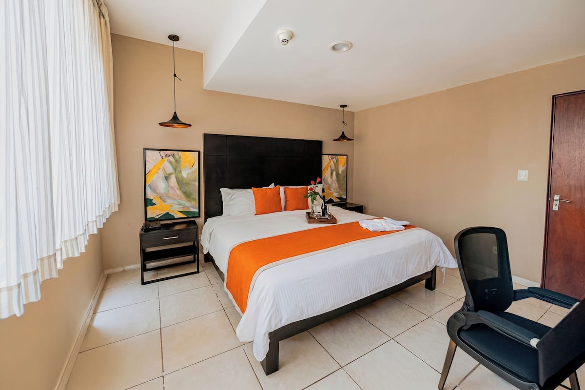 A hotel room with a bed, a chair, a nightstand, and a wall-mounted artwork.