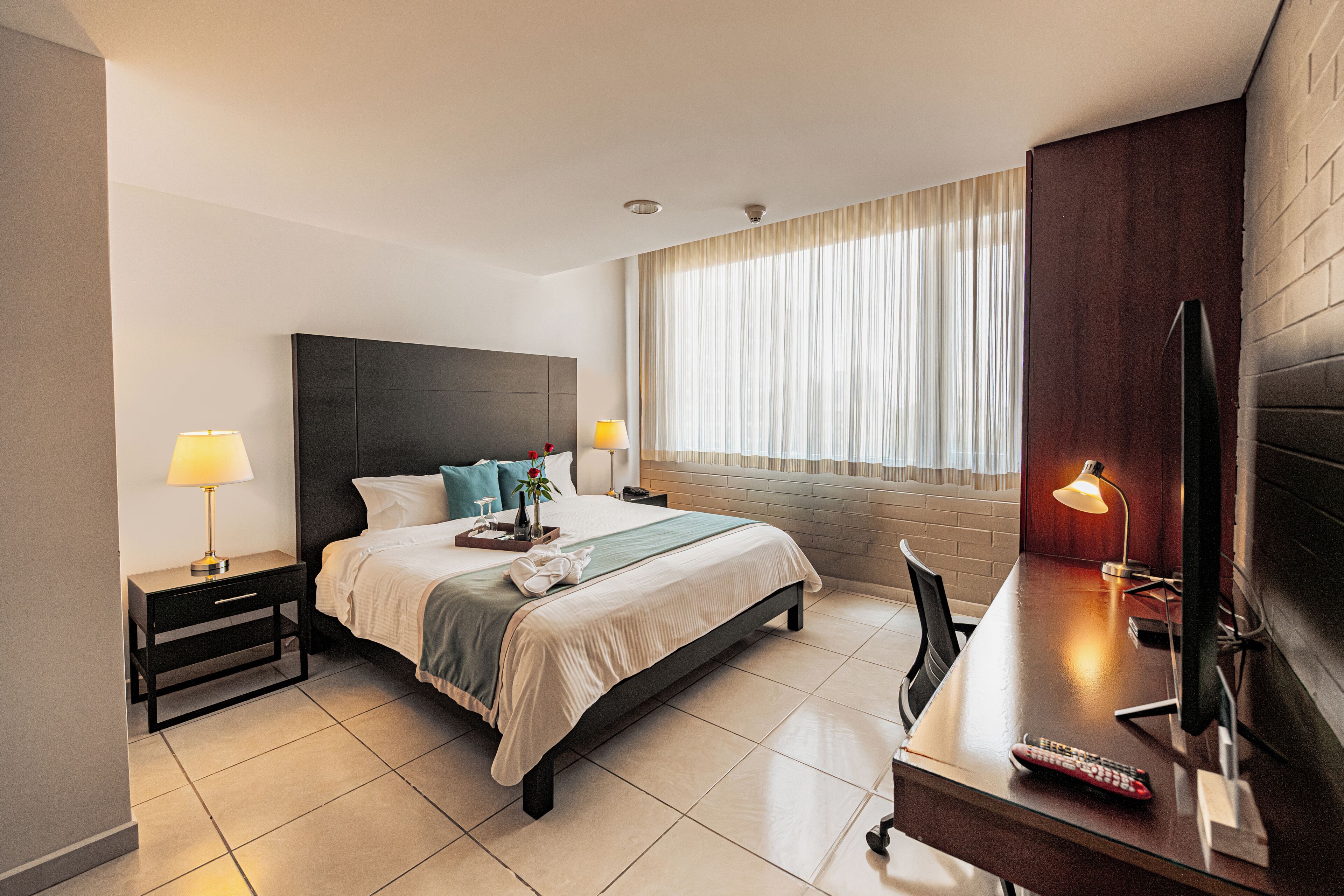 A hotel room with a bed, bedside tables, a desk with a lamp, a TV, and a window with curtains.