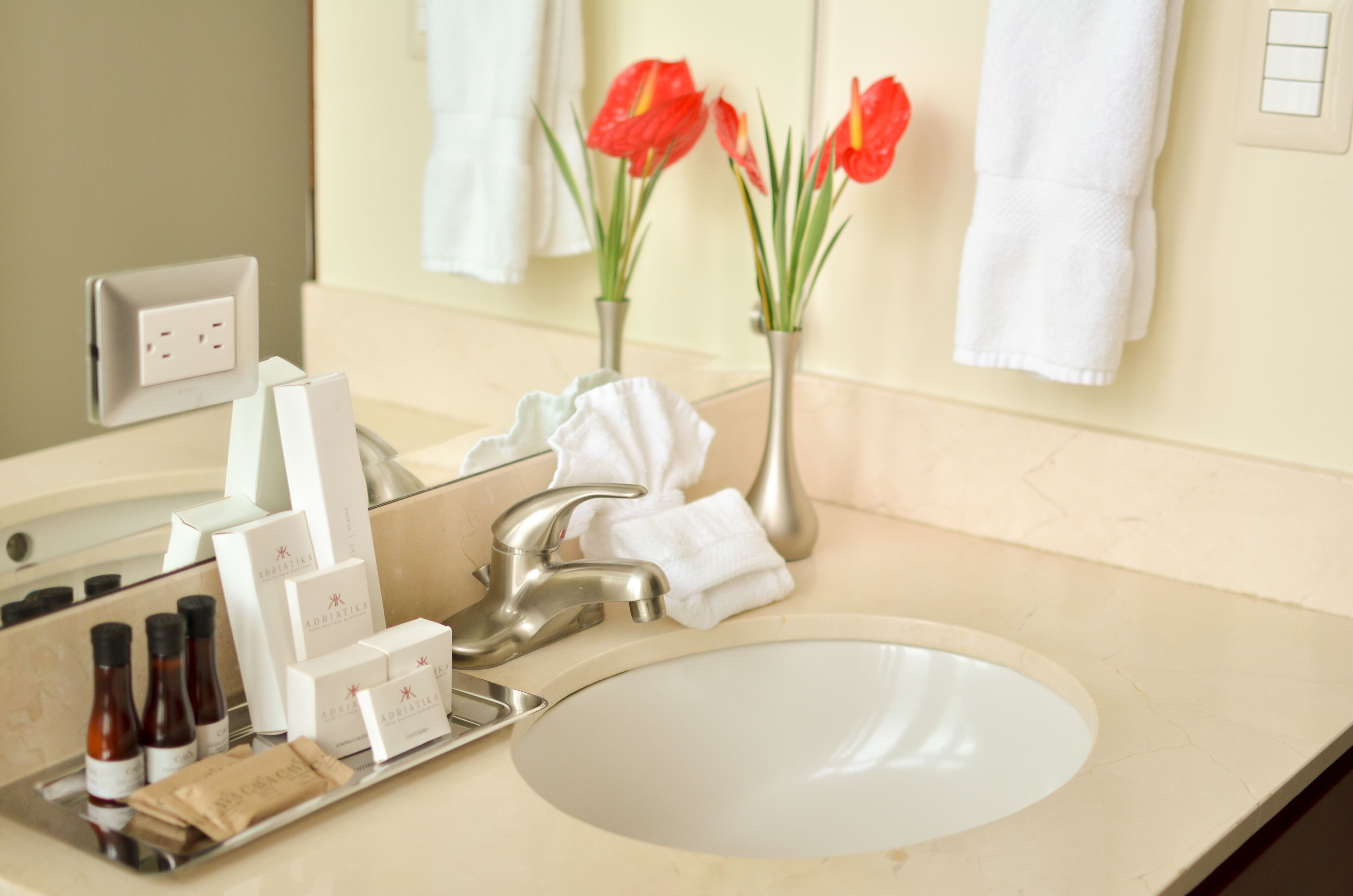 Un baño con lavamanos, espejo, artículos de tocador y flores rojas.