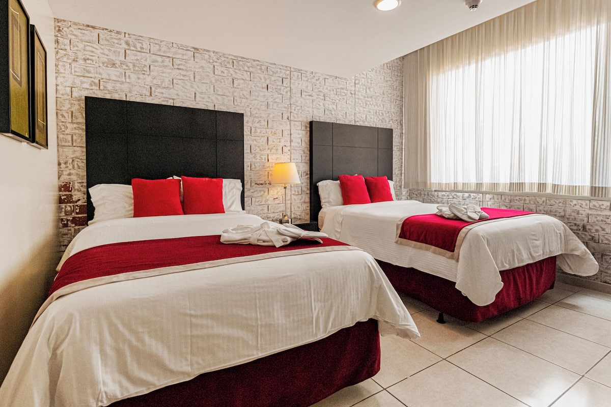 A hotel room with two beds, a brick-patterned wall, a large window with curtains, and a nightstand with a lamp.