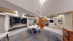 A modern hotel lobby with a central dining area, a reception desk, and a large mirror.