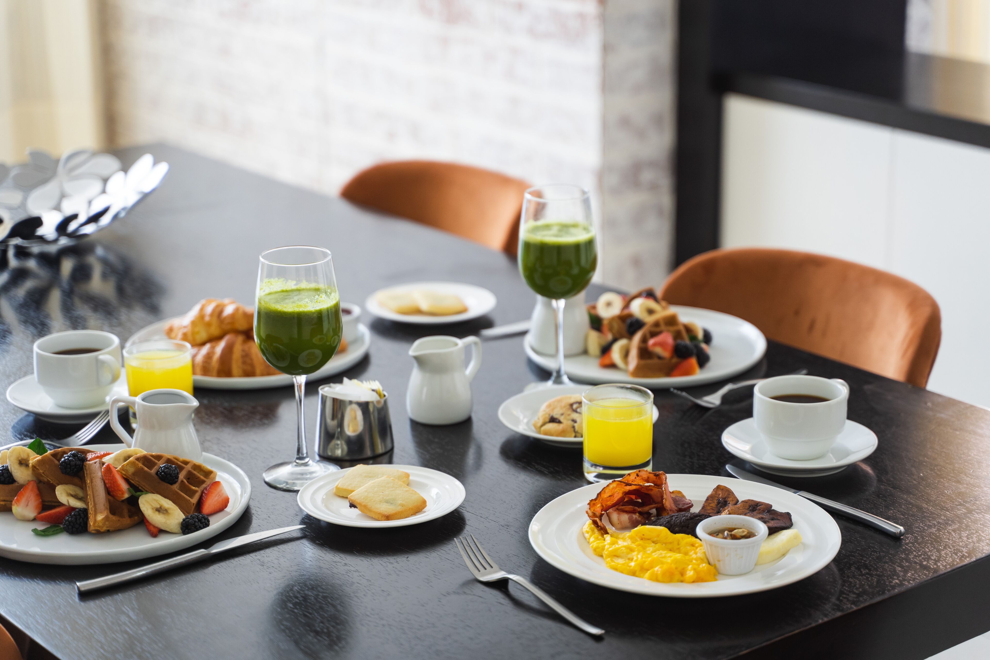 Una mesa de desayuno con diversos platos, como waffles, huevos y tostadas, junto con tazas de café y vasos de jugo de naranja.
