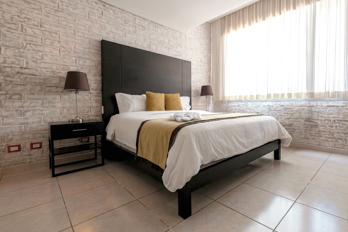 A modern hotel room with a brick-patterned wall, a large bed with white bedding and a brown throw, two bedside lamps, and a nightstand with a lamp.