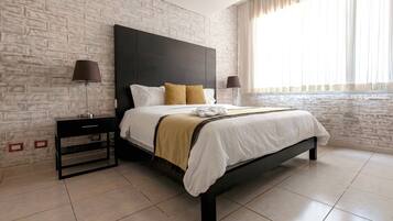 Una habitación de hotel moderna con pared de ladrillos, una cama grande con ropa blanca y un plaid marrón, dos lámparas de noche y una mesita de noche con una lámpara.