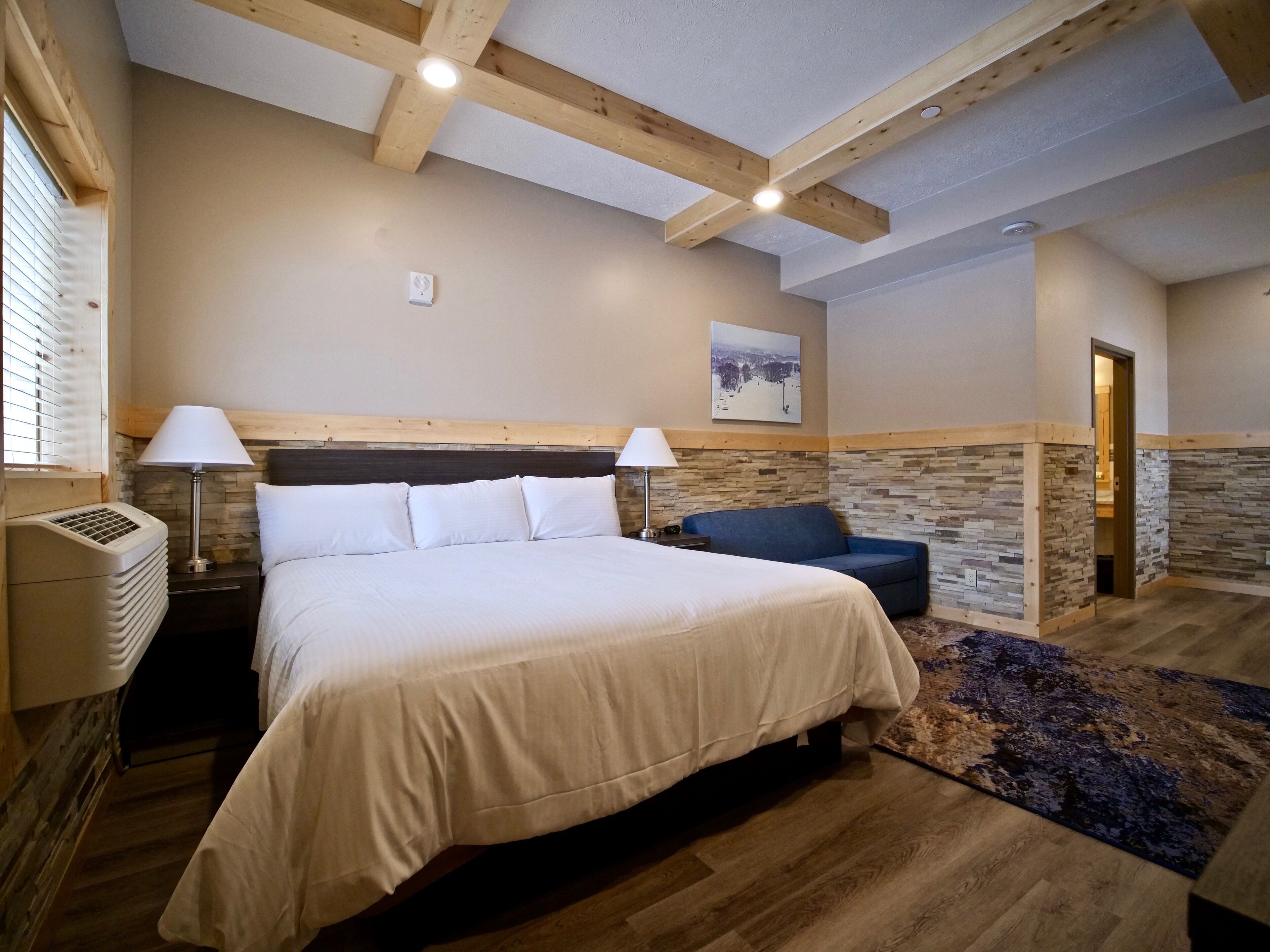 Un dormitorio con una cama grande, dos lámparas de noche, una pared decorativa de piedra, una silla azul y un techo de madera.