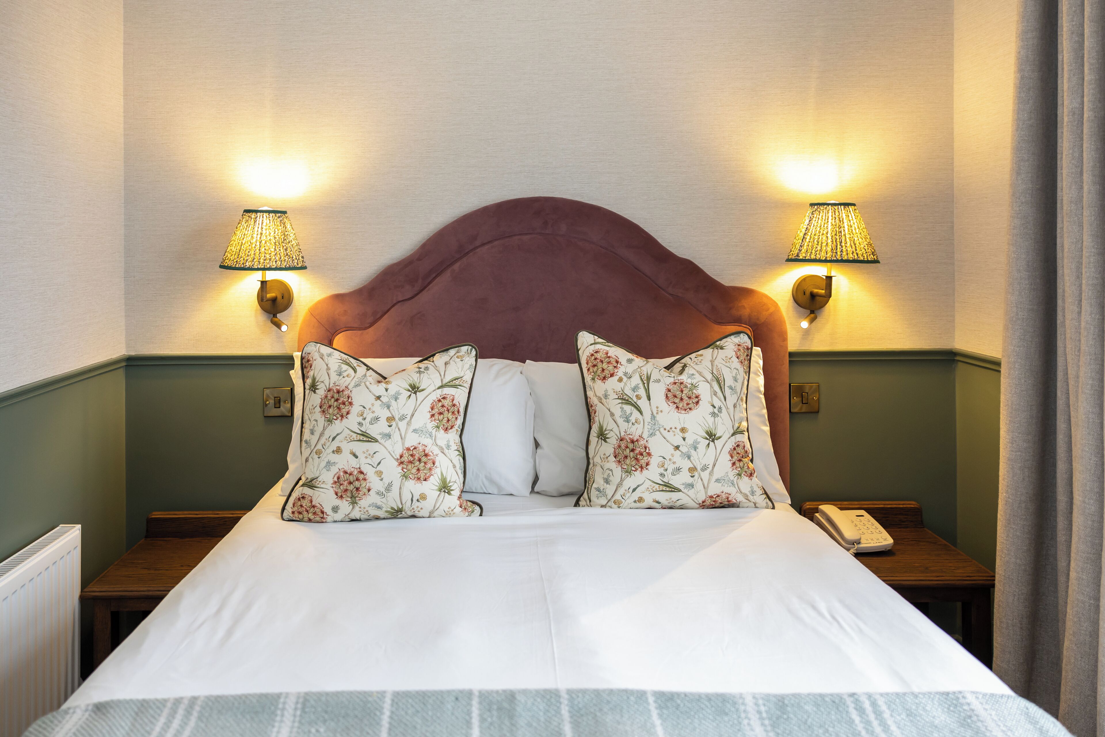 Una cama bien hecha con almohadones de estampado floral, dos lámparas de noche y un teléfono sobre una mesita de madera.