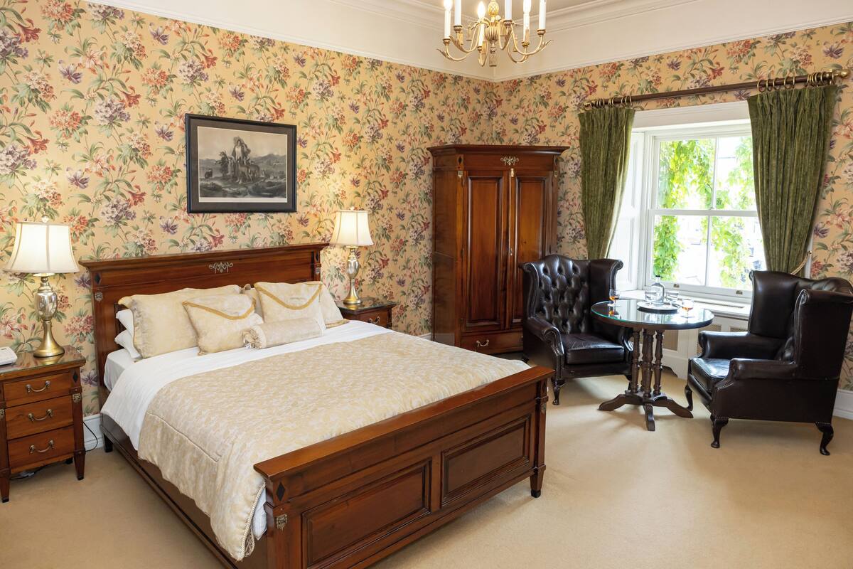 Quarto com uma cama grande, guarda-roupa de madeira, poltrona de couro, mesa redonda pequena e janela com cortinas.