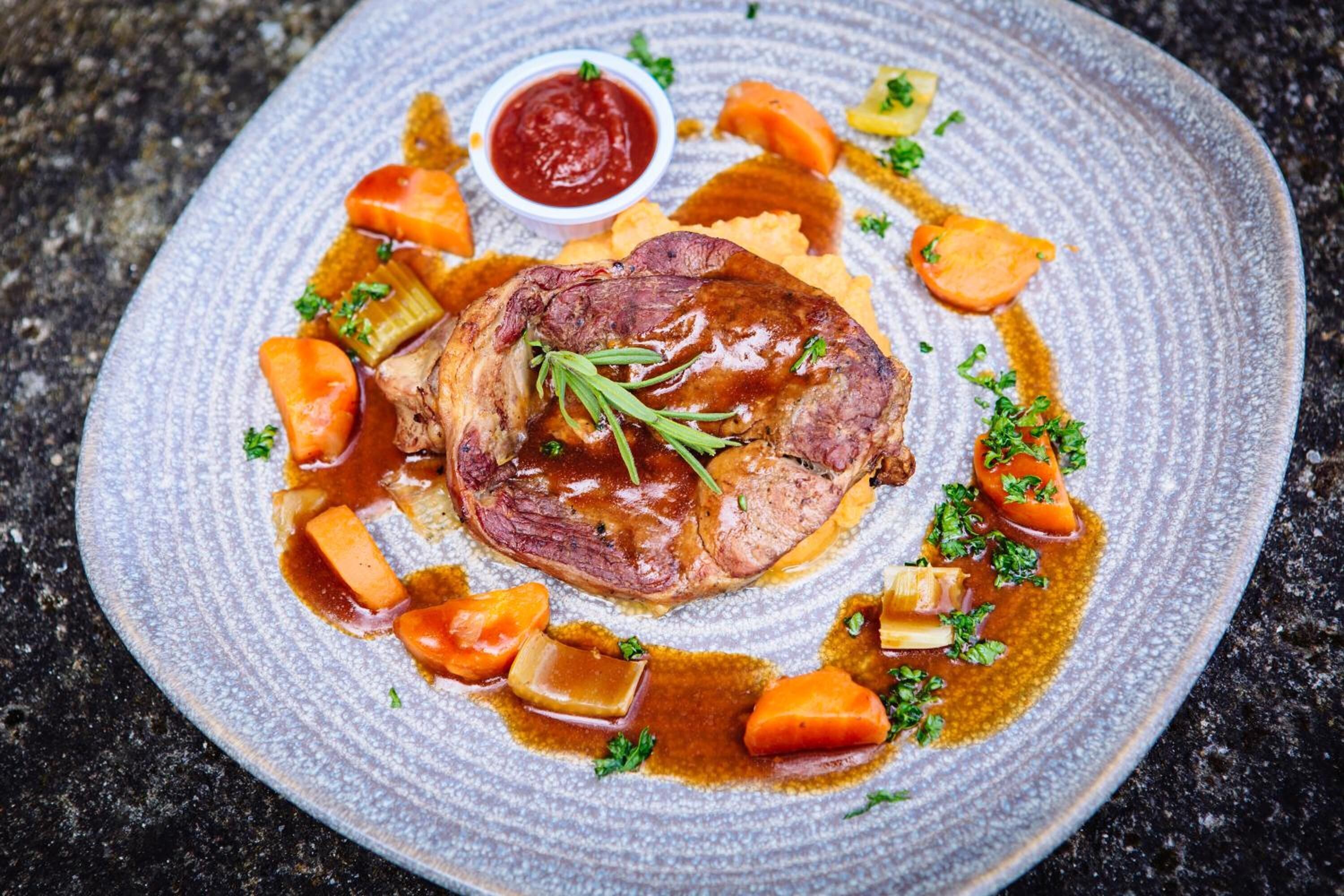 A plate of food with a steak, carrots, and a small bowl of sauce.