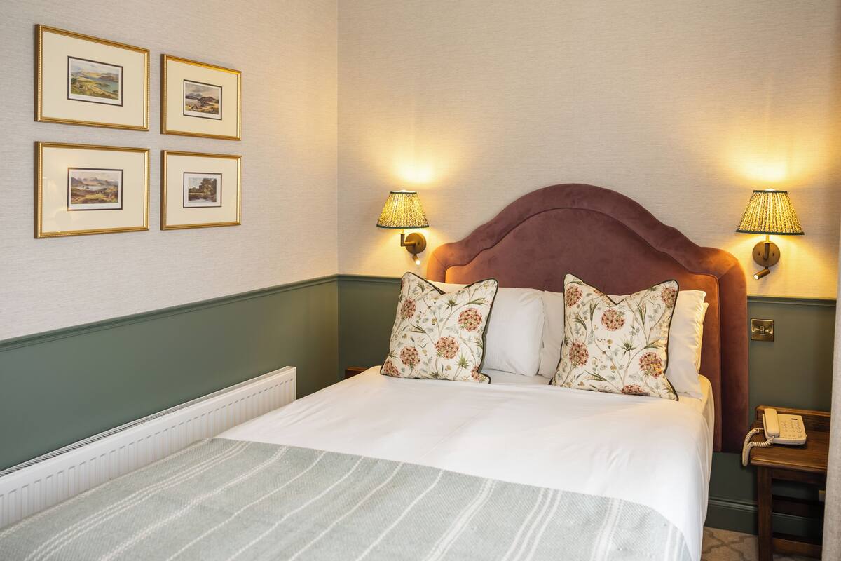 Uma cama bem arrumada com almofadas florais, cabeceira e duas luminárias fixadas na parede.