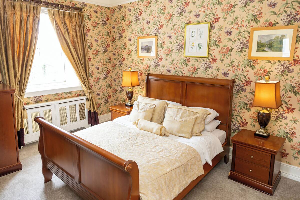 Quarto com cama de madeira, duas mesas de cabeceira com abajures, papel de parede floral e quadros nas paredes.