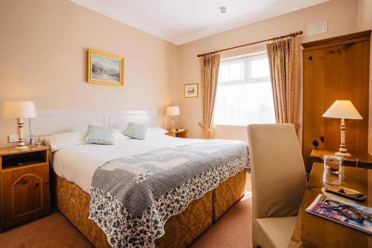 Quarto de hotel com cama, duas mesas de cabeceira, uma cadeira, escrivaninha, janela com cortinas e um quadro na parede.
