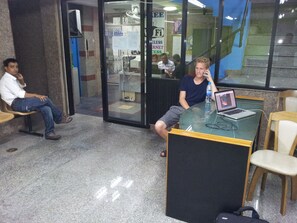 A waiting area with a glass partition, a laptop on a table, and a person sitting on a bench. - Centaur Inn (Bangkok)