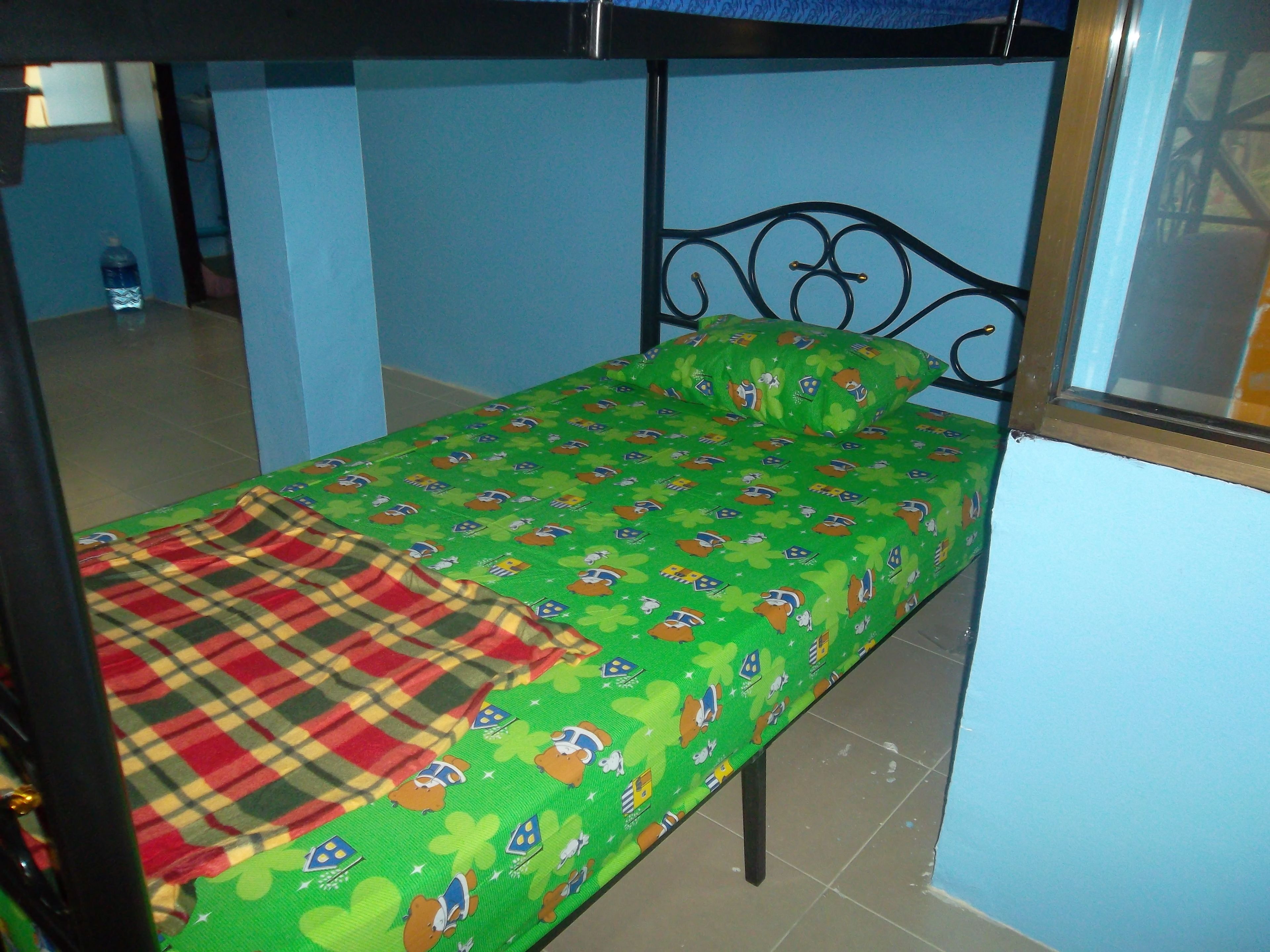 A single bed with a green bedspread featuring cartoon characters, a plaid blanket, and a metal headboard.