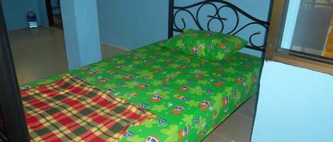 A single bed with a green bedspread featuring cartoon characters, a plaid blanket, and a metal headboard.