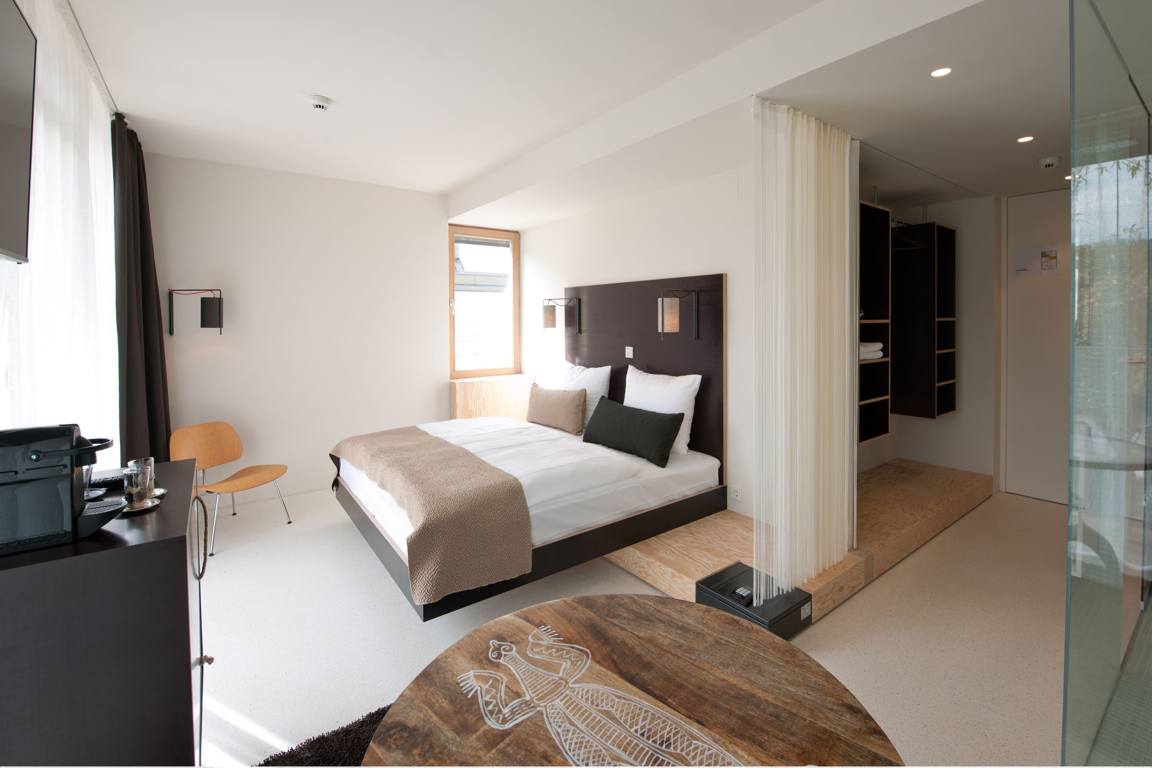 A modern hotel room with a large bed, a desk, a chair, and a glass-enclosed shower area.