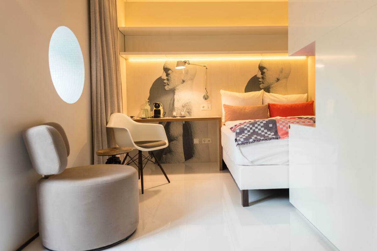 A modern hotel room with a white bed, a desk, a chair, and a large mirror.