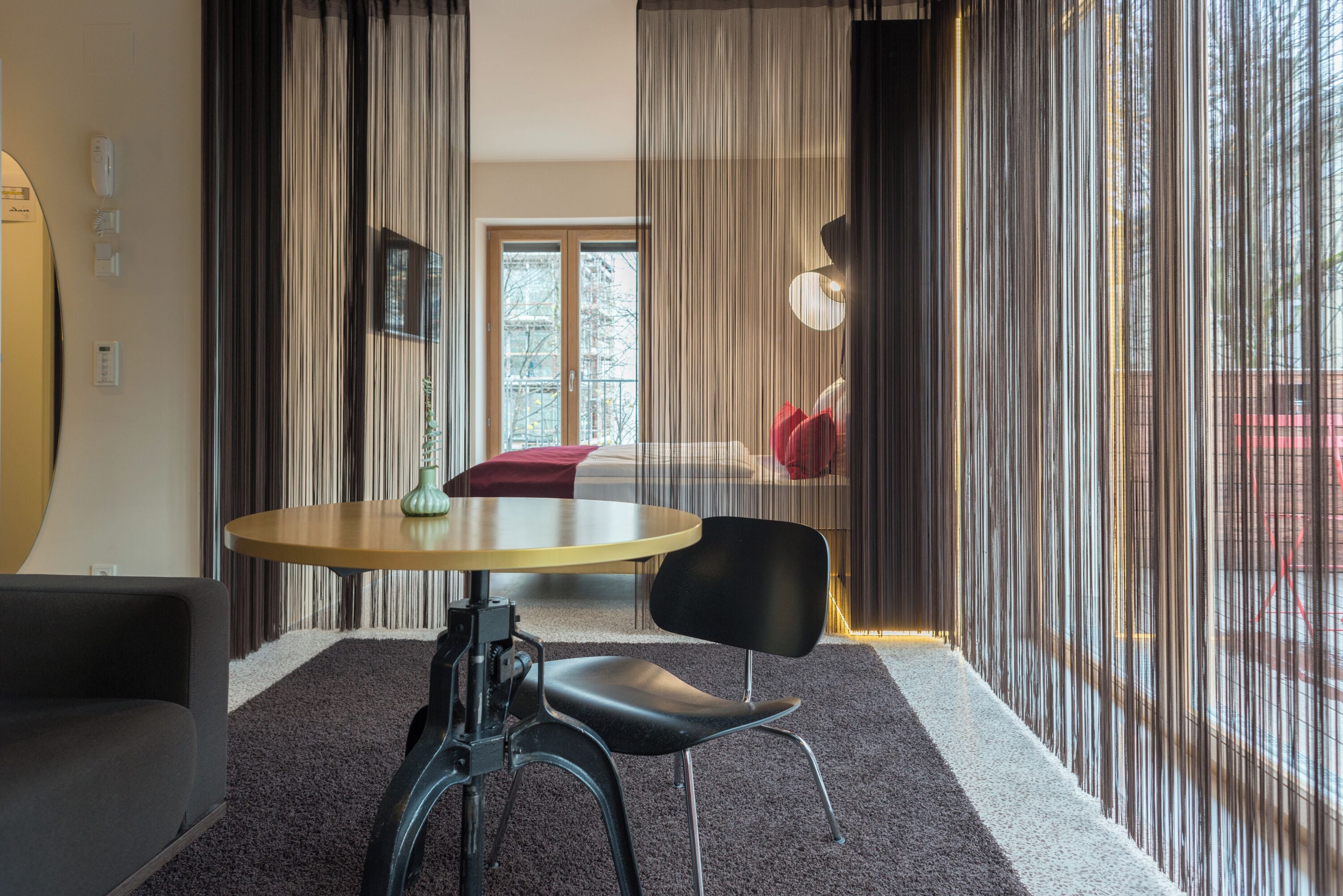 Une pièce moderne avec une table ronde, une chaise noire et un canapé. On y trouve une fenêtre avec des rideaux, une porte et une vue sur l’extérieur.