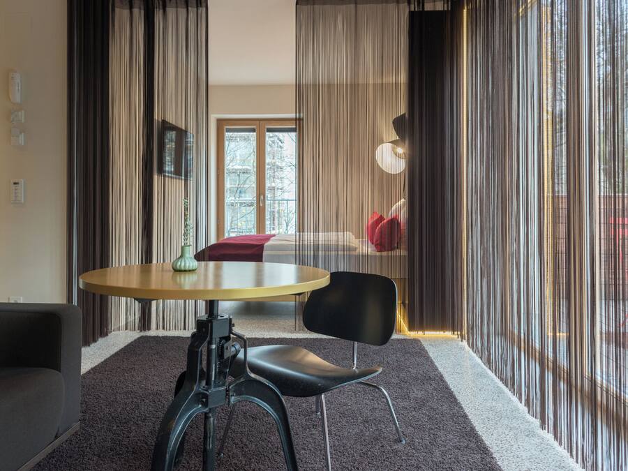A modern room with a round table, a black chair, and a sofa. There is a window with curtains, a door, and a view of the outside.