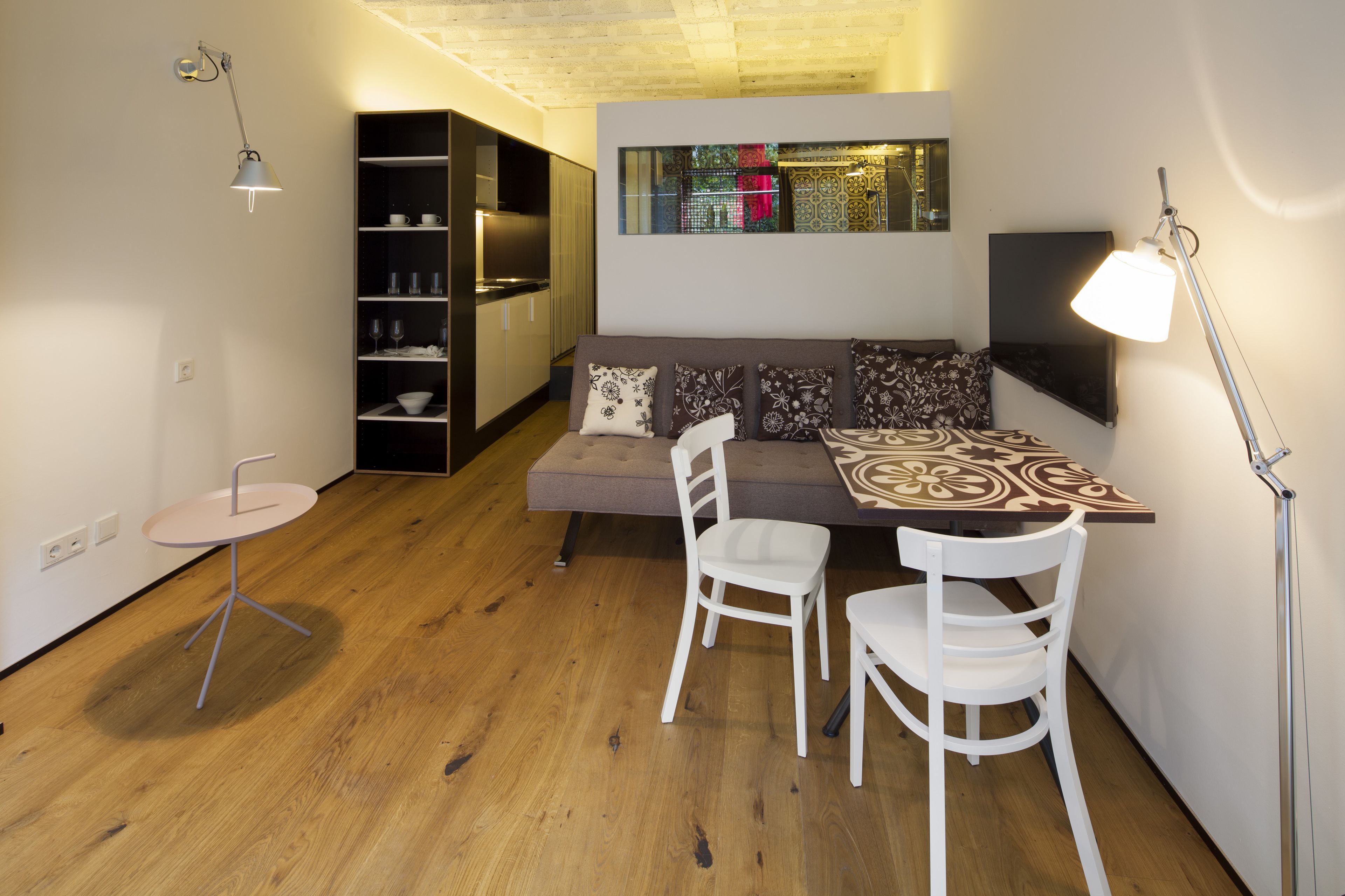Een gezellig compacte ruimte met een bank, twee witte stoelen, een klein rond tafeltje, een boekenkast en een vloerlamp.