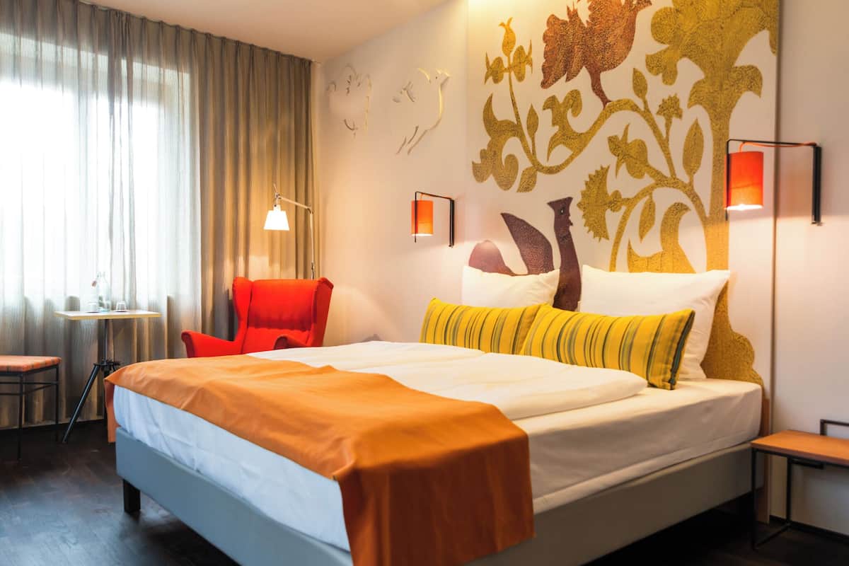 A hotel room with a large bed, a red armchair, a small table with a lamp, and a decorative wall mural.