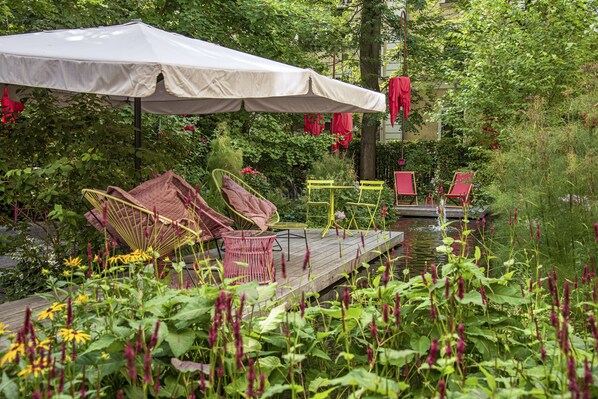 A garden patio with outdoor furniture, a white umbrella, and a wooden deck over a pond. - NALA individuellhotel (Innsbruck)