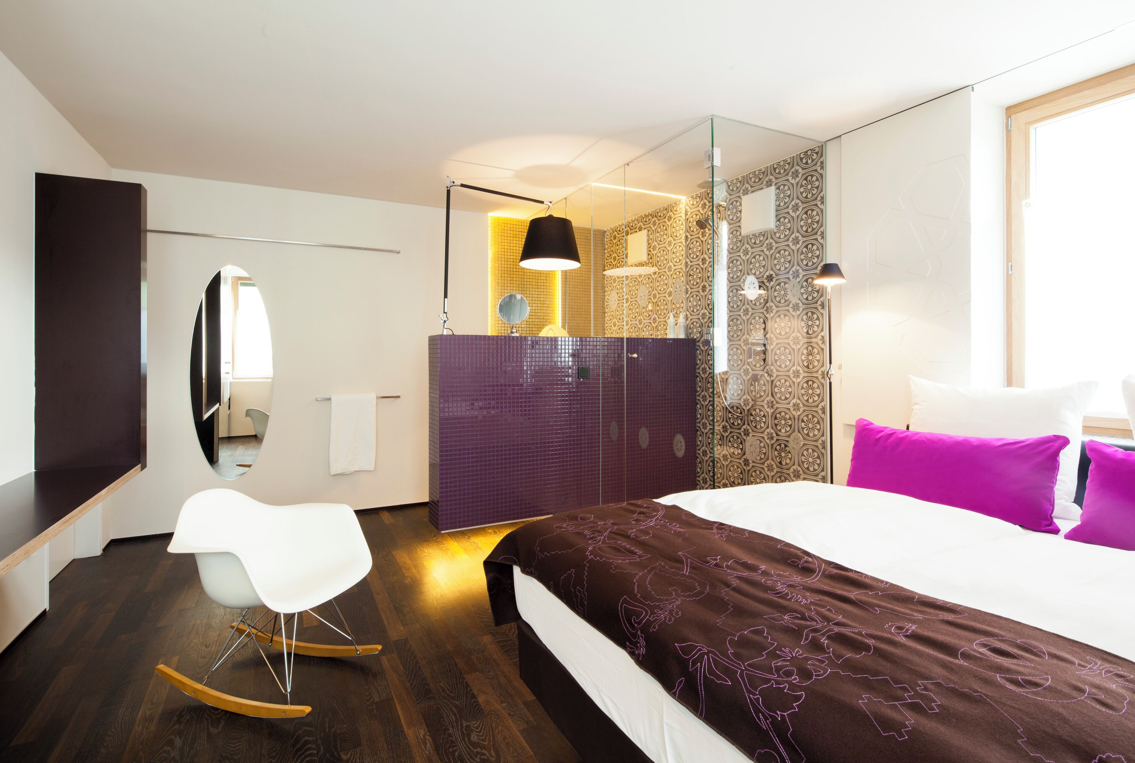 A modern hotel room with a large bed, a white rocking chair, a bathroom with a glass partition, and a patterned wall.