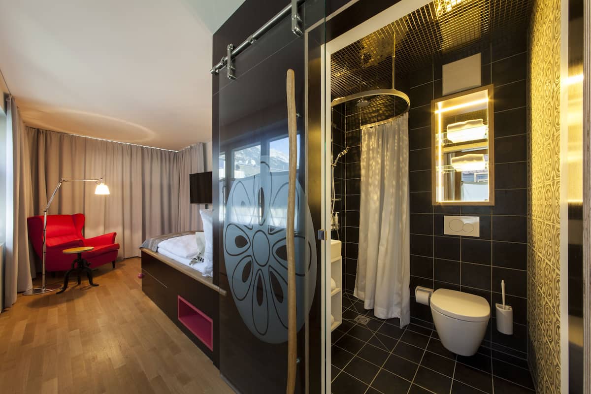 A hotel room with a bed, a red chair, a desk, a shower, and a toilet.
