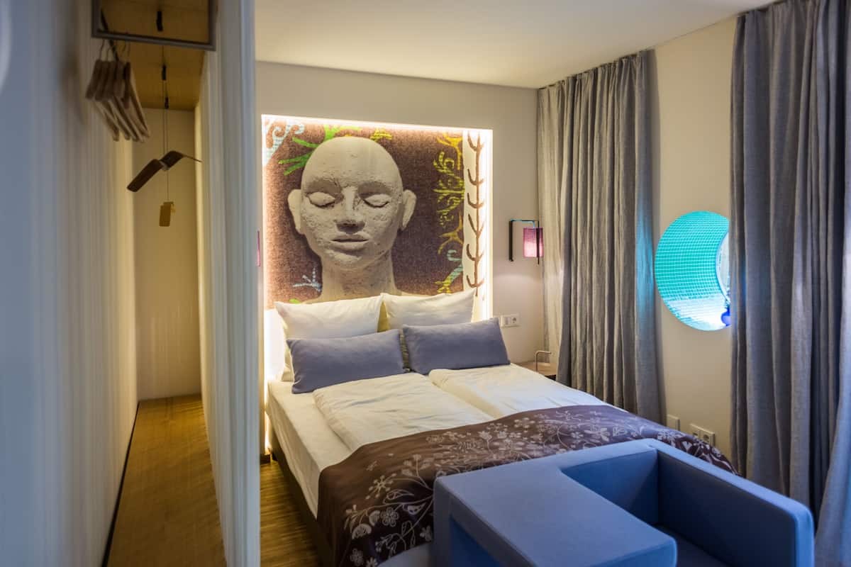 A modern hotel room with a bed, a large wall art piece, a side table, and a window with curtains.