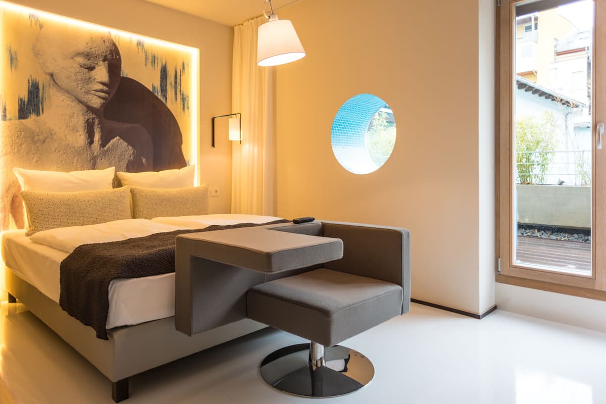 A modern bedroom with a bed, a side table, a round window, and a wall-mounted lamp.