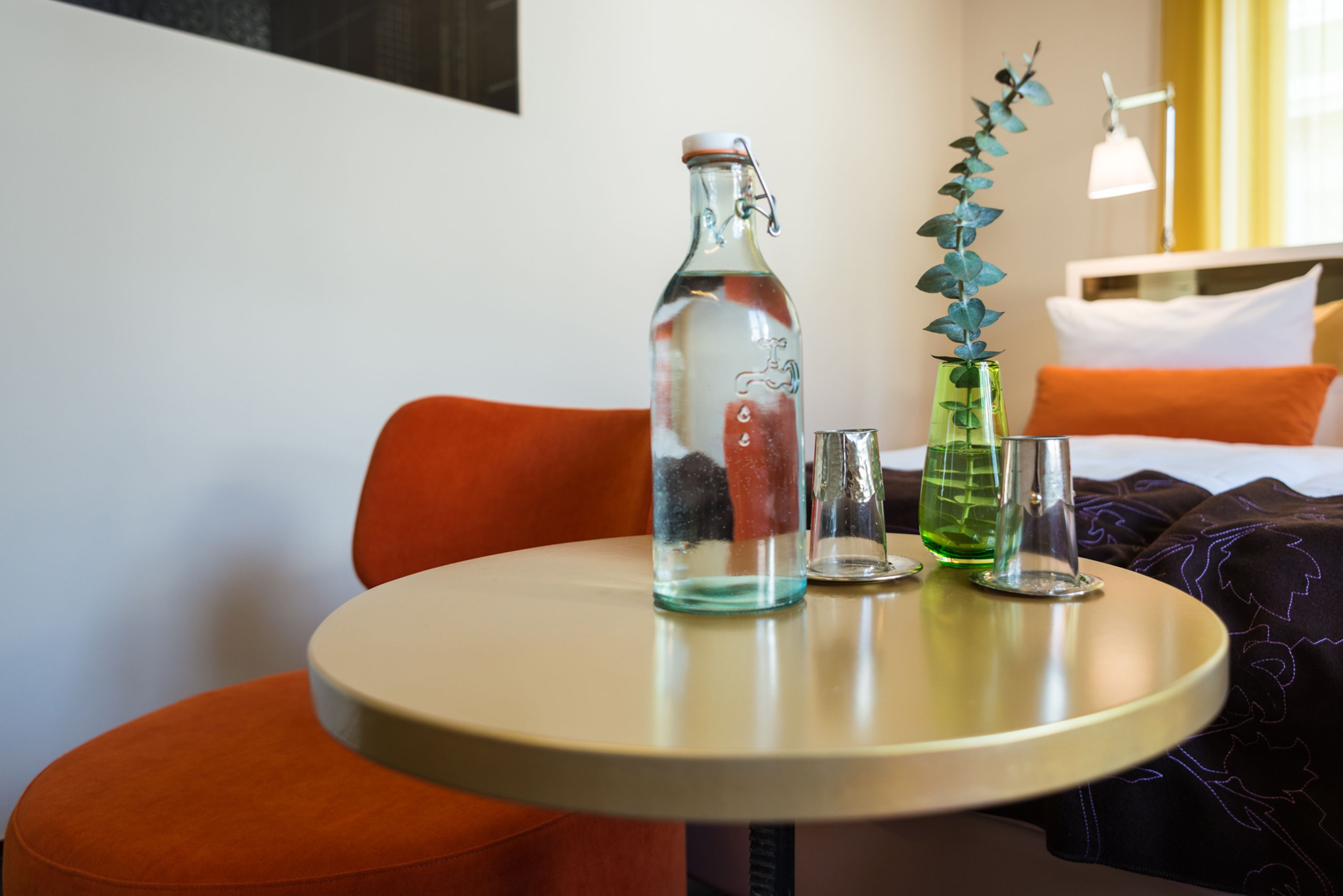 Een kleine ronde tafel met een fles en twee glaasjes, een bloempot en een bed met oranje kussens op de achtergrond.