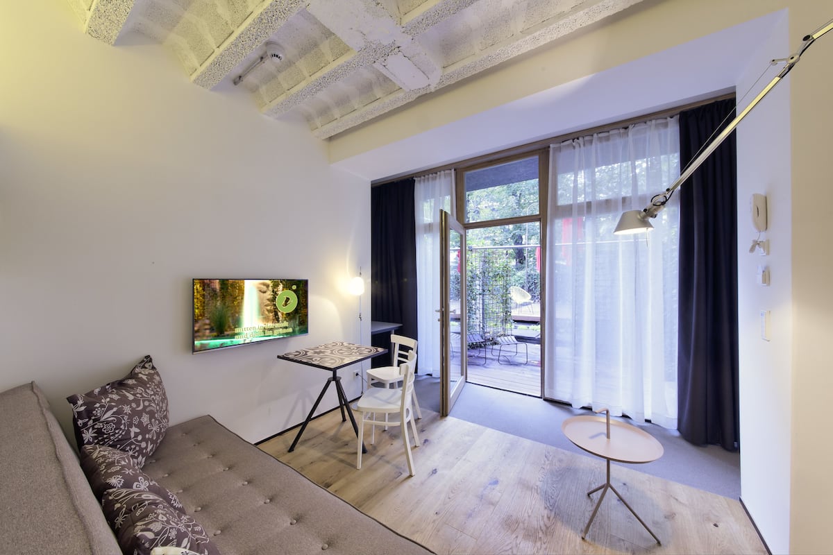 A modern living room with a sofa, a small table, and a chair. There is a large window with curtains, a ceiling with exposed beams, and a wall-mounted TV.