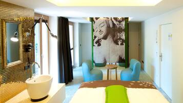 Quarto de hotel com uma cama grande, banheiro com pia e espelho, duas cadeiras e uma parede com uma obra de arte de grandes dimensões.