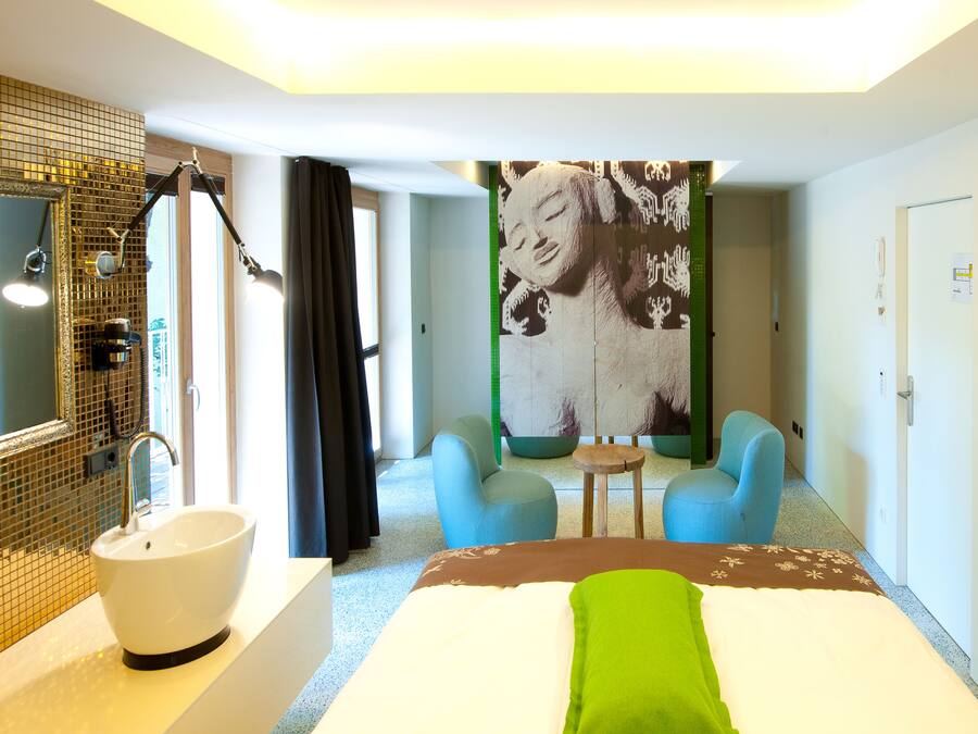 Quarto de hotel com uma cama grande, banheiro com pia e espelho, duas cadeiras e uma parede com uma obra de arte de grandes dimensões.