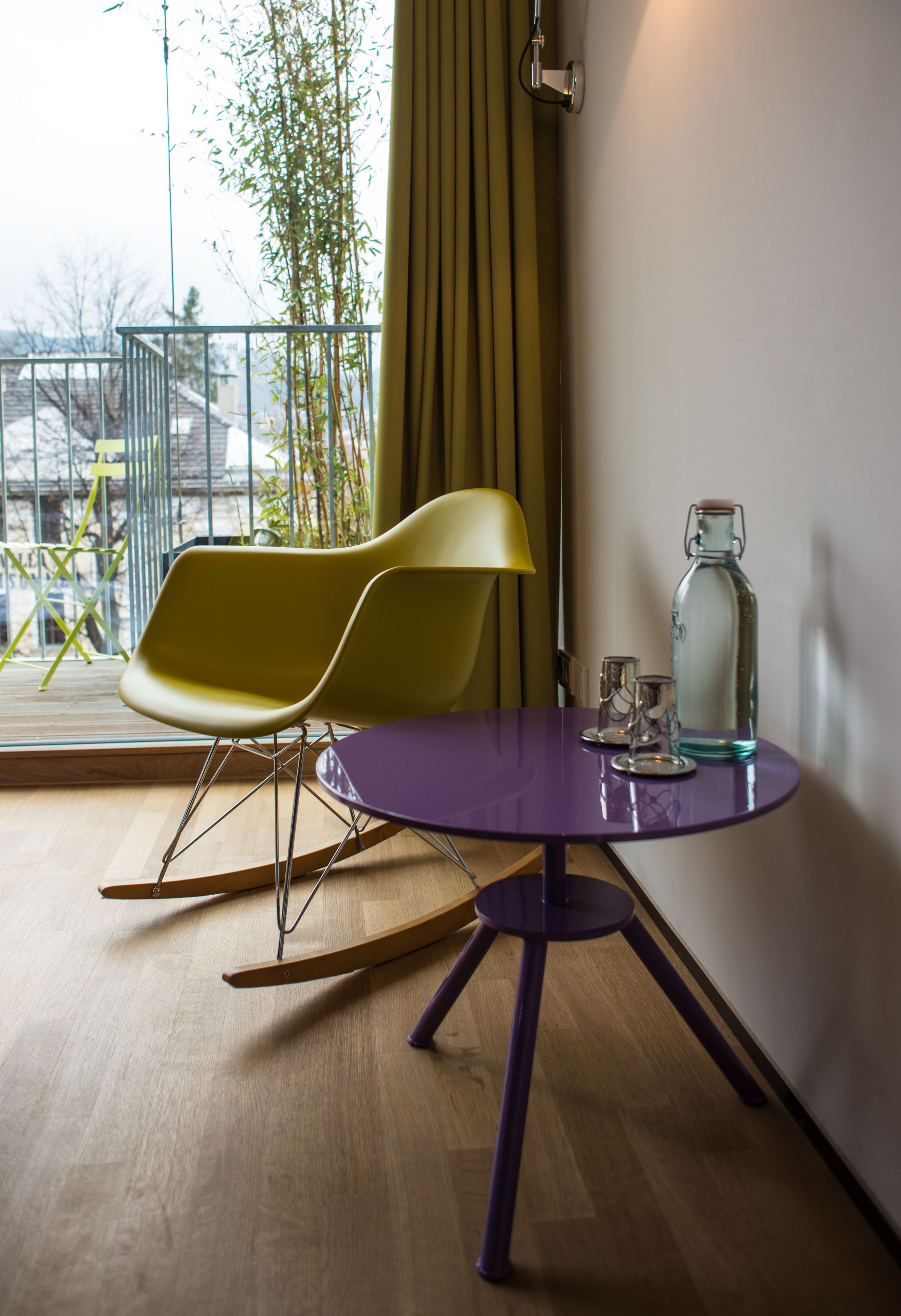 A room with a purple table, two yellow chairs, a glass bottle, and a window with a view of a building and trees.