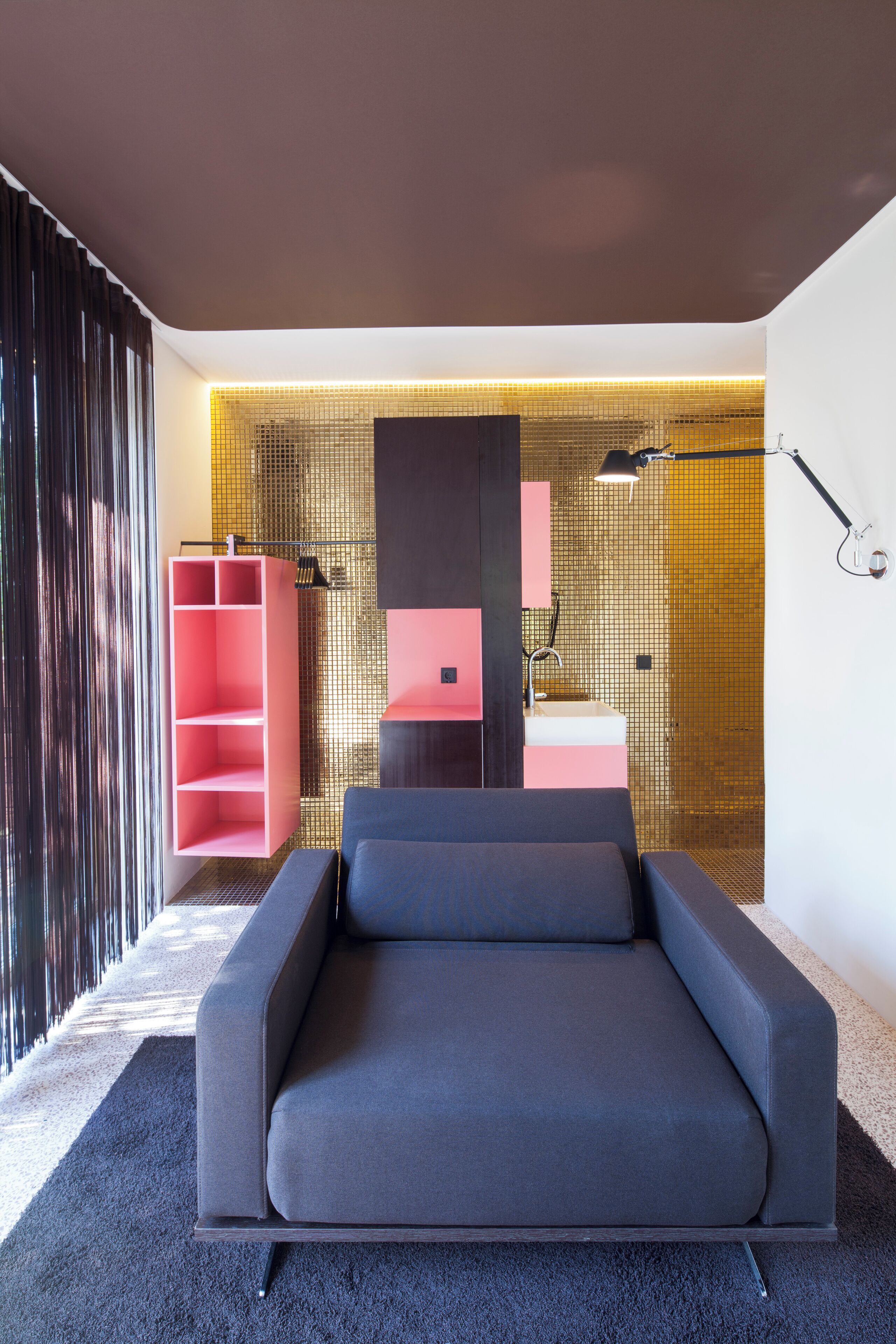 A modern bedroom with a grey sofa, a pink shelving unit, and a bathroom with a tiled wall.