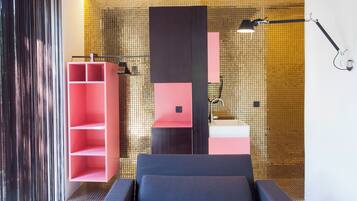 A modern bedroom with a grey sofa, a pink shelving unit, and a bathroom with a tiled wall.