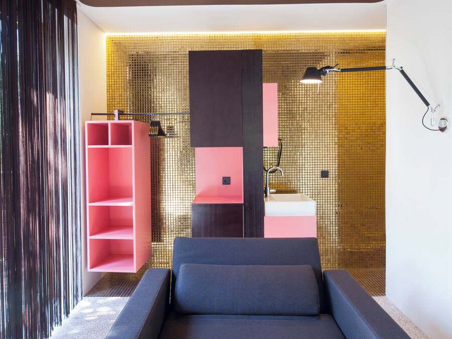 A modern bedroom with a grey sofa, a pink shelving unit, and a bathroom with a tiled wall.
