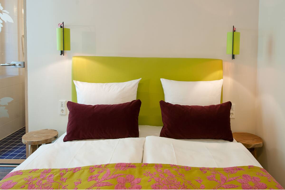 A hotel room with a bed, two pillows, a bedside table, and a wall-mounted lamp.