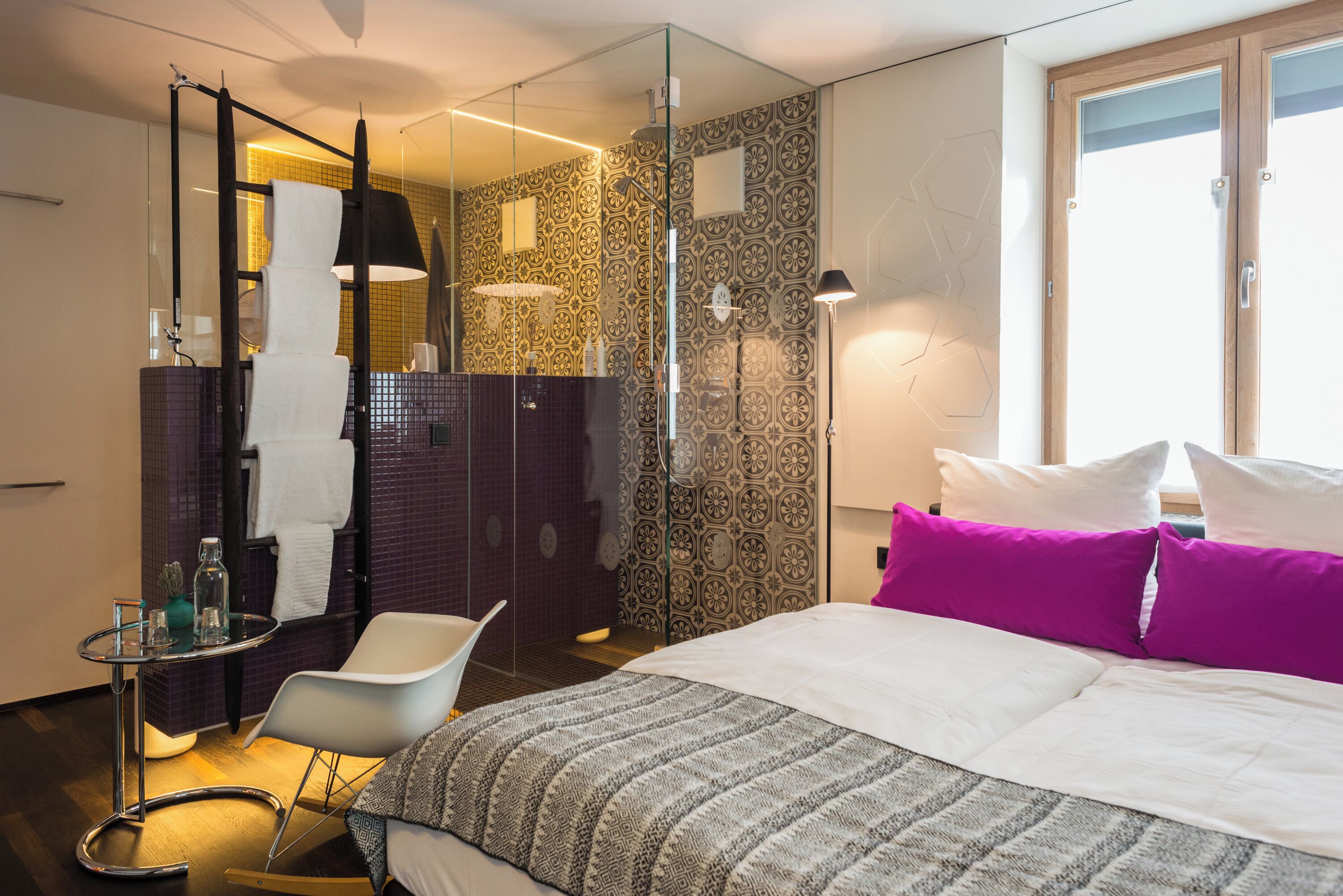 A modern hotel room with a large bed, a glass shower enclosure, a small table with a chair, and a large window.